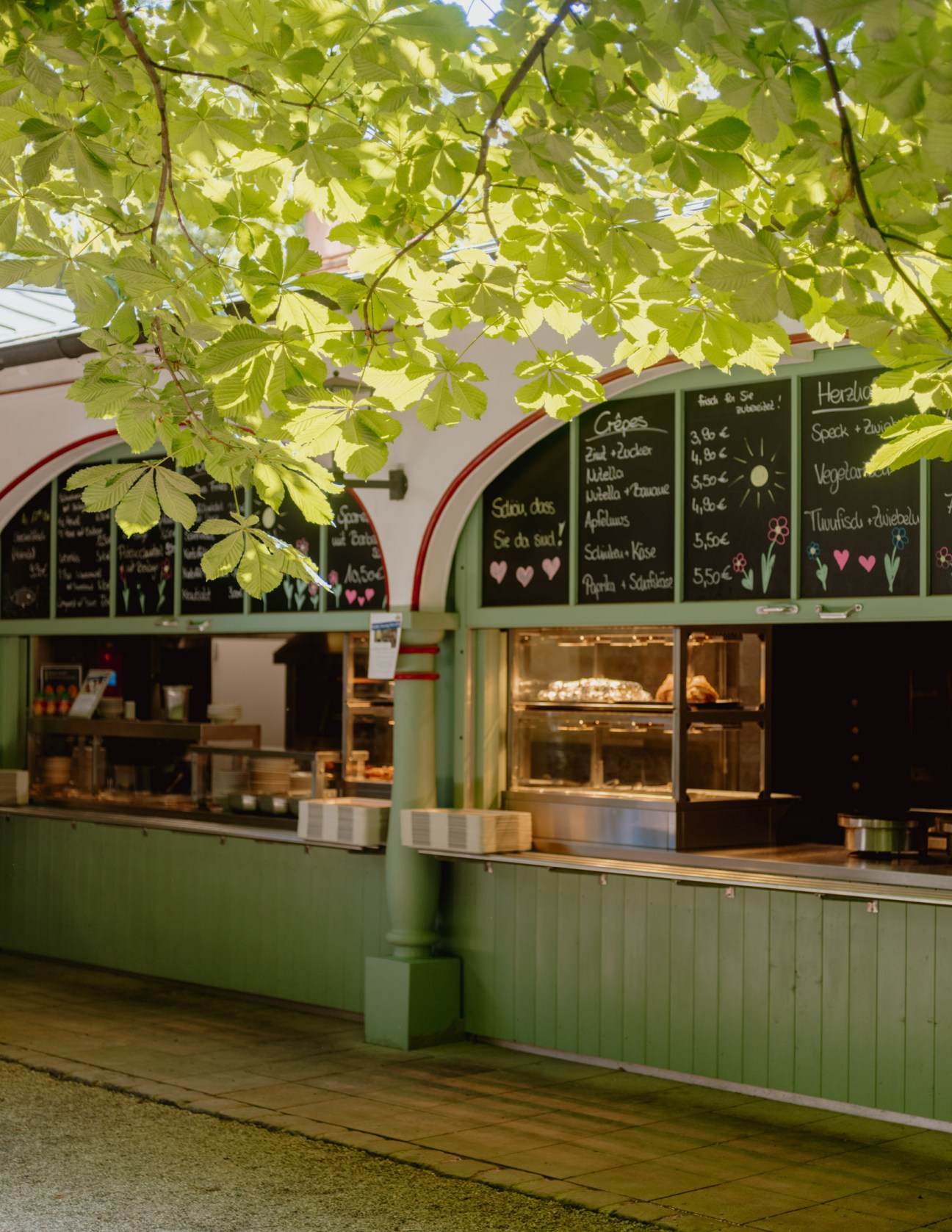 A green food stall with blackboard menus under dappled horse chestnut tree leaves at a Bavarian beer garden.