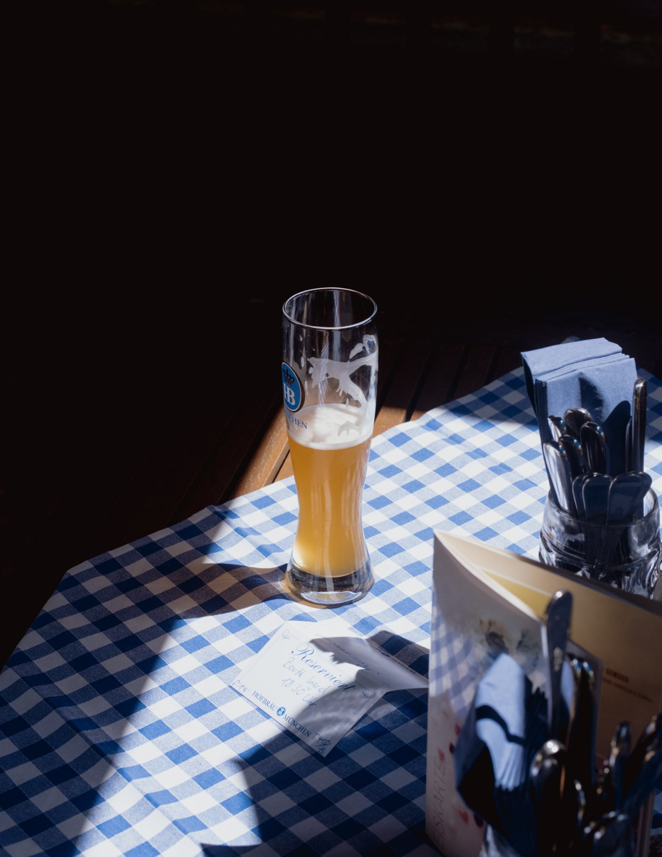 A glass of beer on a blue and white checkered tablecloth with utensils and napkins.