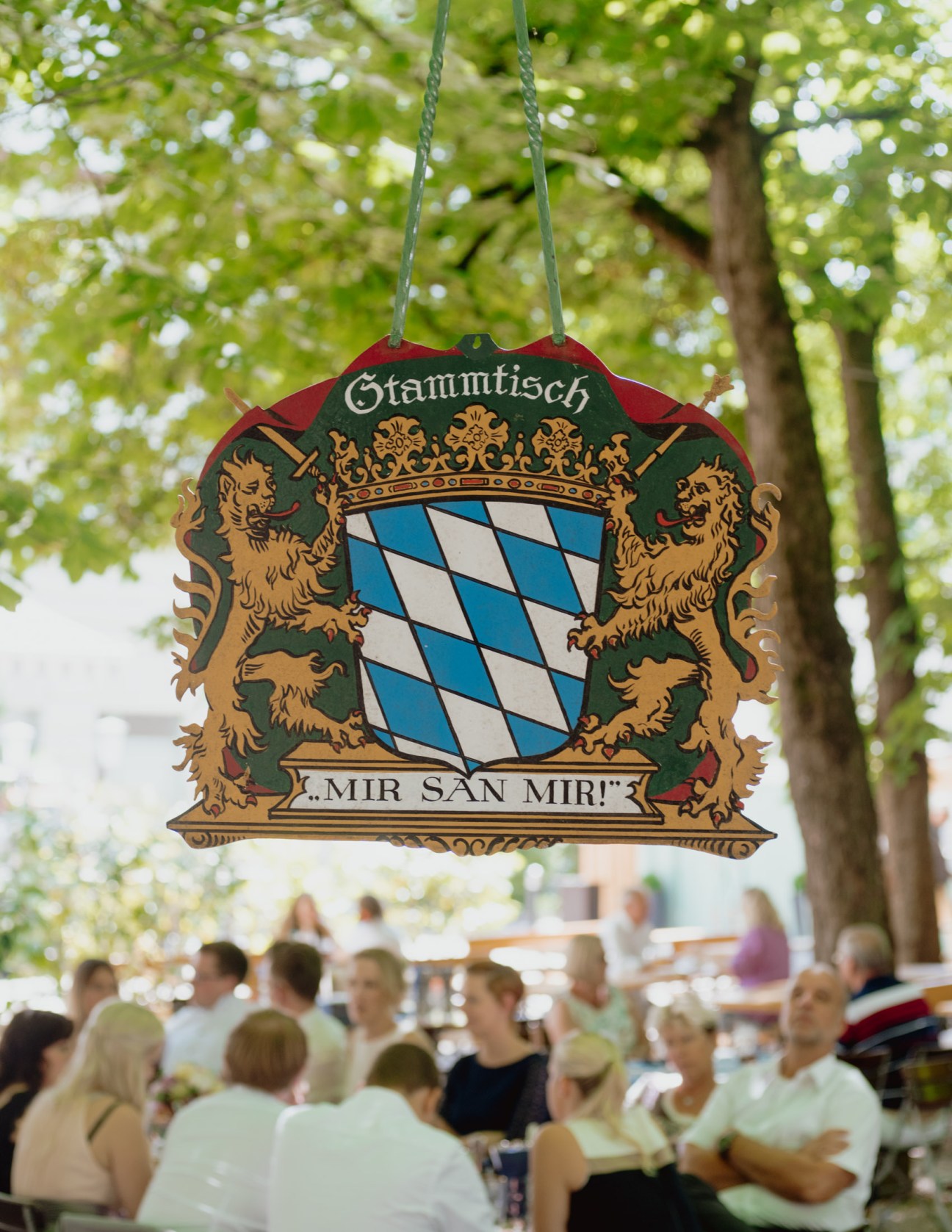 Ornate wooden sign hanging from chains reading 'Stammtisch' with Bavarian coat of arms and lions, suspended above beer garden patrons beneath chestnut trees.