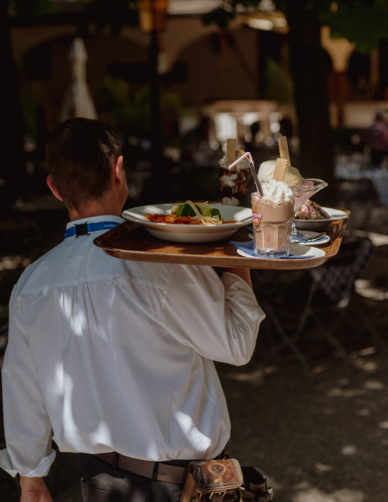 Waiter in white shirt carries tray with food and drinks at a beer garden.