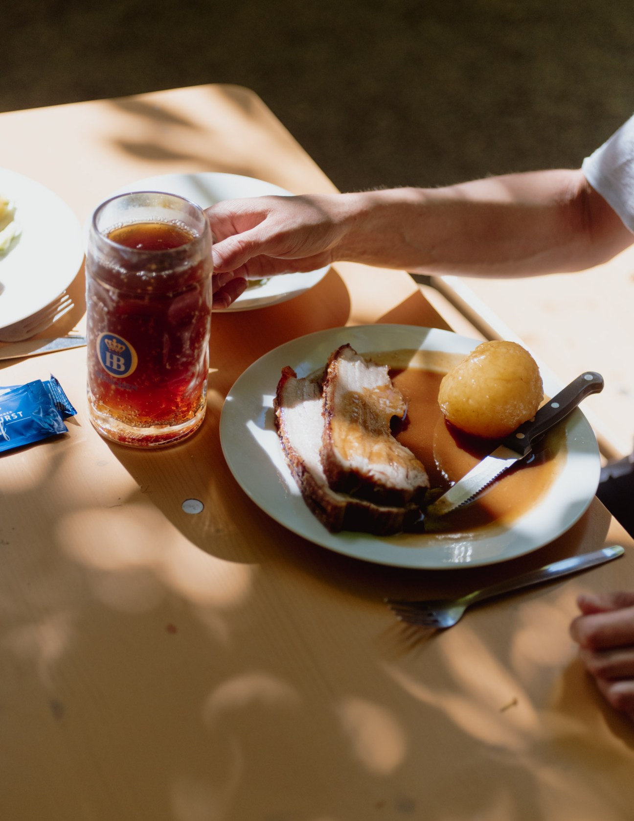 Hand holding a beer glass next to a plate of roasted meat and potatoes at a beer garden table.