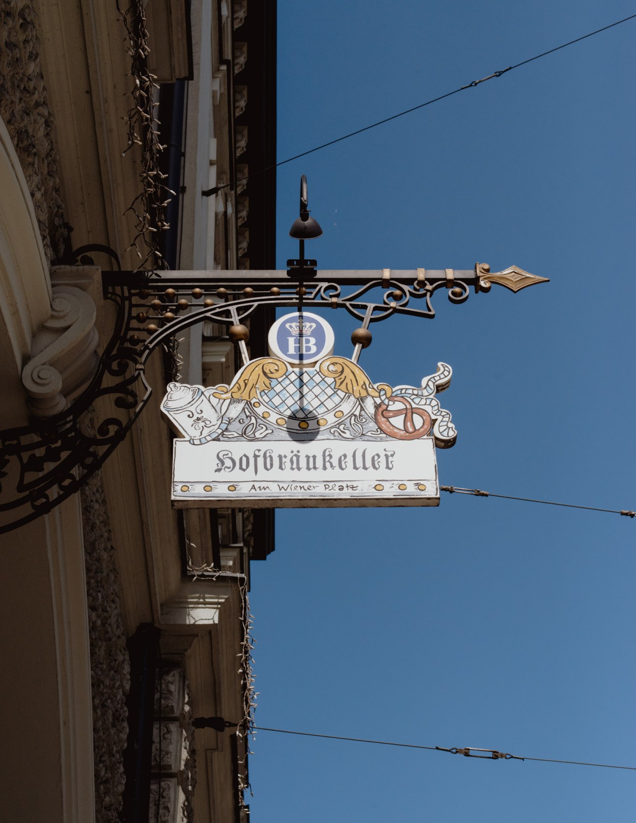 Ornate wrought iron sign for Hofbräukeller beer hall with HB brewery logo against clear blue sky.