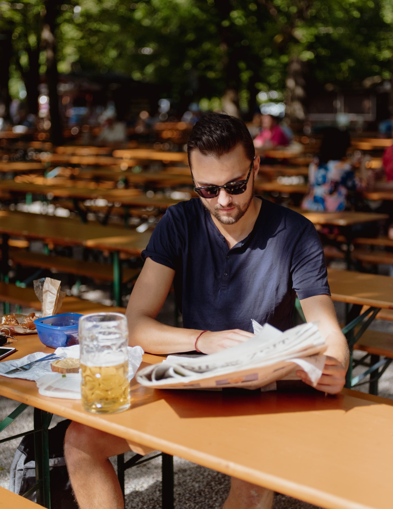Man reading newspaper while drinking beer at outdoor beer garden table under trees.