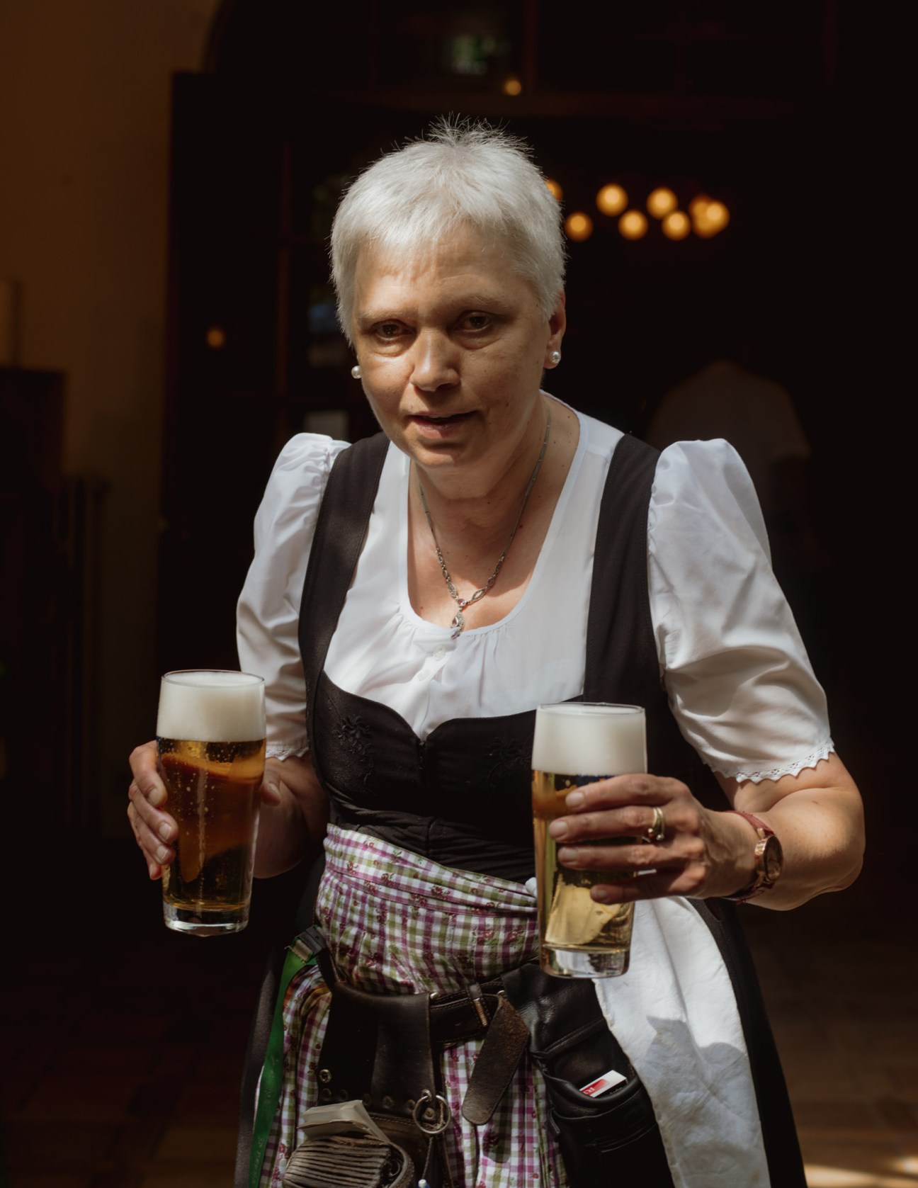 A waitress in traditional Dirndl dress carries two full beer glasses in a Munich beer garden.