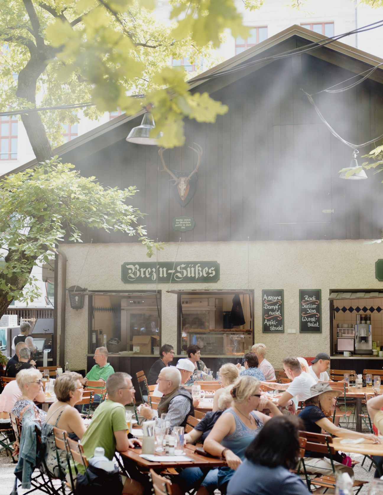 Crowded Munich beer garden with patrons seated at wooden tables, a service counter, and trees overhead.