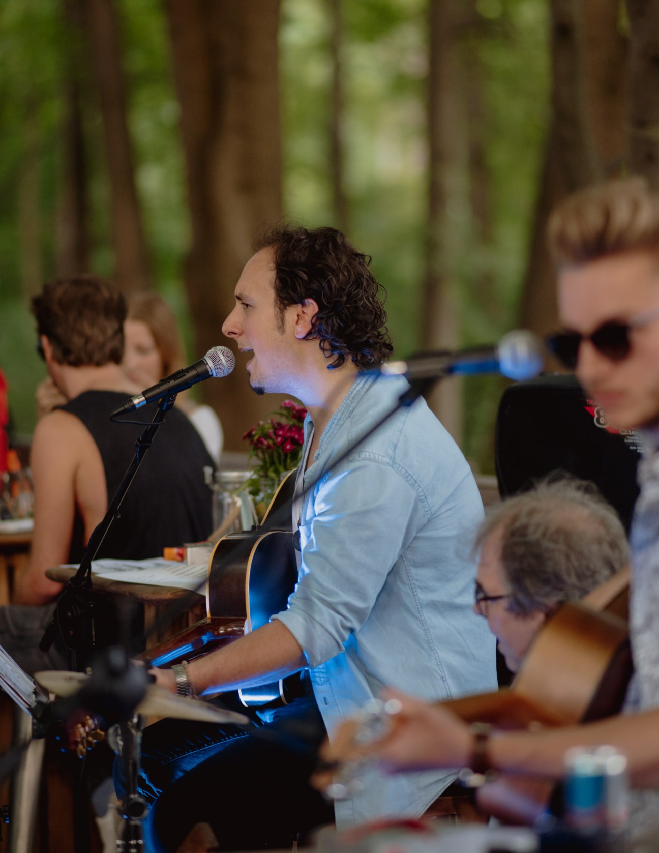 Musician performing with guitar and microphone at an outdoor beer garden event surrounded by trees.