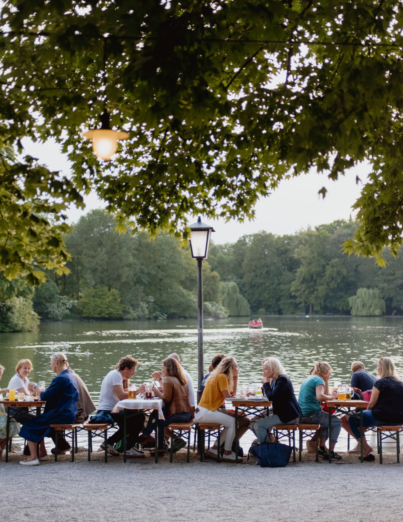People dining at outdoor tables by a lake surrounded by trees, with a rowing boat on the water in the background.