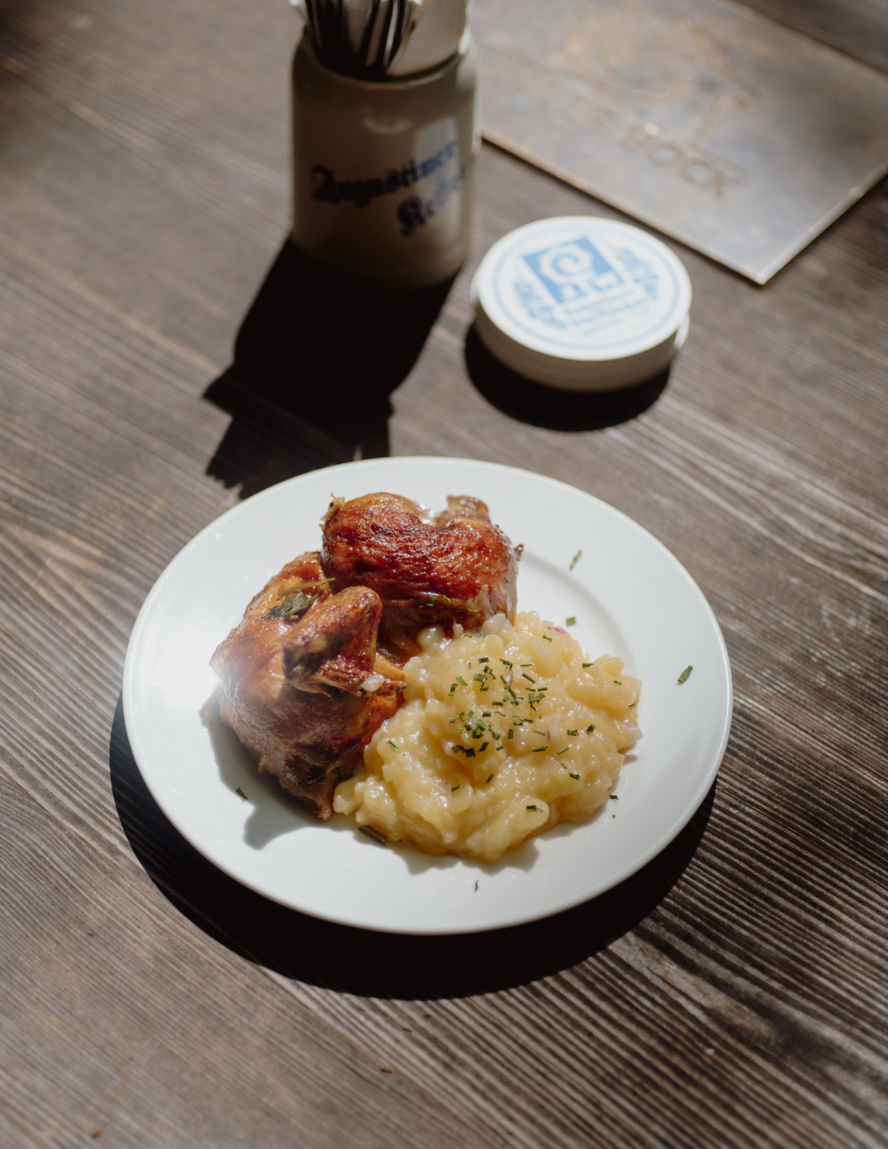 Roasted chicken with potato salad on a white plate next to a beer stein and coaster on a wooden table.