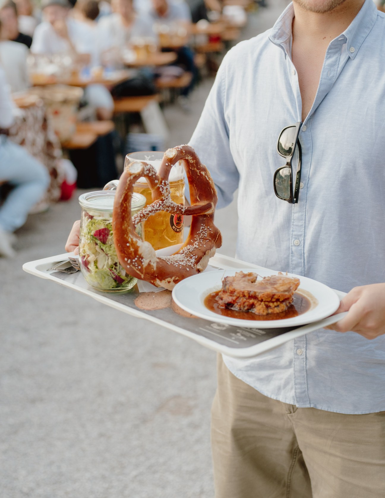 A man in a light blue shirt holds a tray with a pretzel, beer, salad, and food at a Bavarian beer garden.