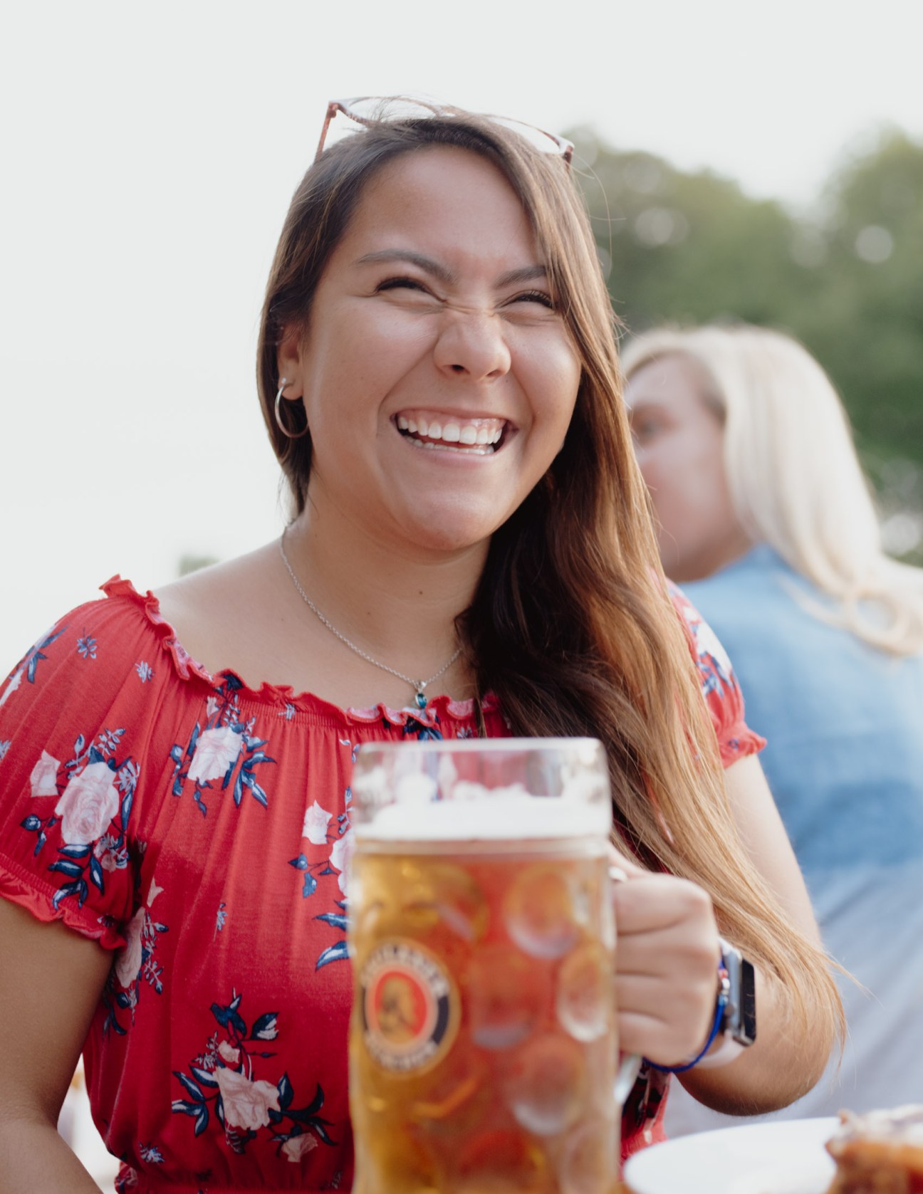Woman in red floral dress smiling while holding a large glass beer stein at a beer garden.