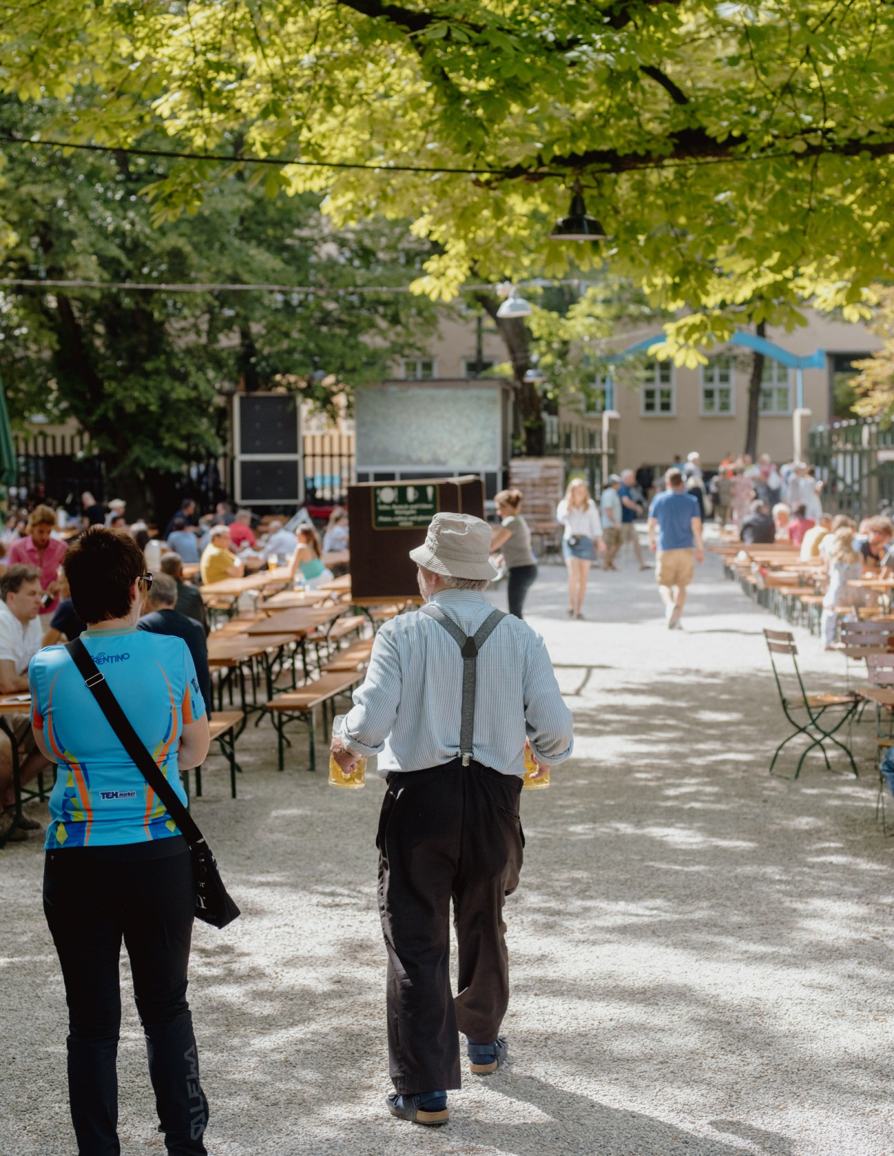 Two people walking through a busy Bavarian beer garden with tables, guests, and trees overhead.