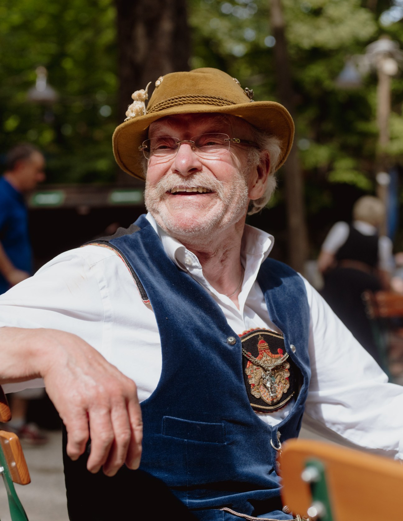Older man wearing traditional Bavarian lederhosen, vest, and hat smiles at a beer garden.
