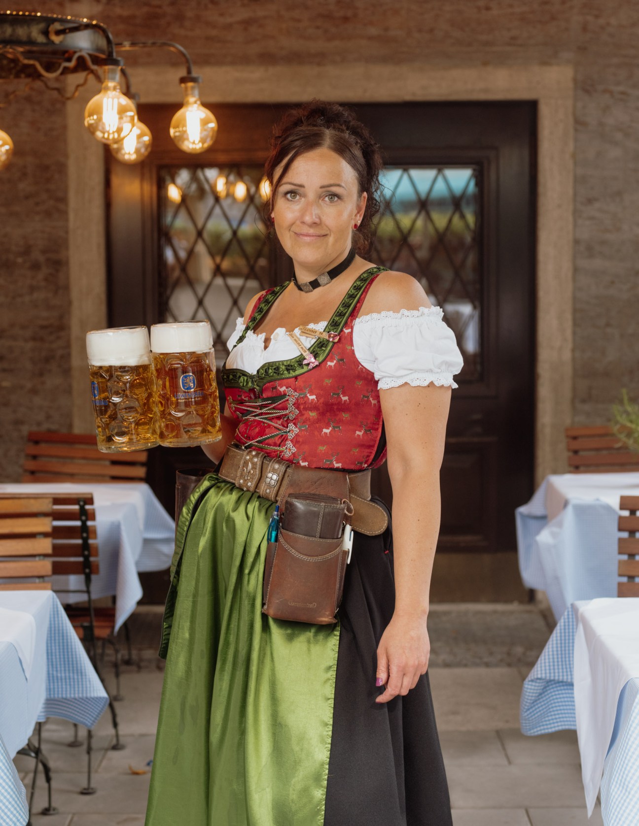 Bavarian waitress in traditional Dirndl dress carrying two beer steins indoors.