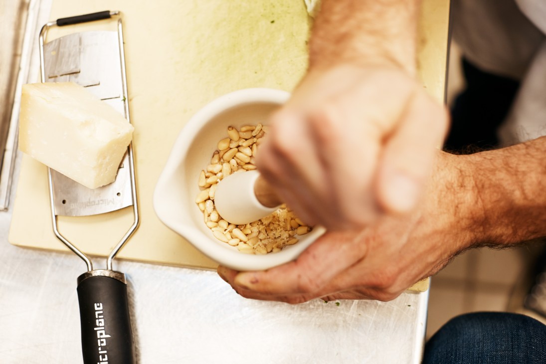 Hands pounding pine nuts in a white mortar and pestle, with shaved cheese and grater visible.