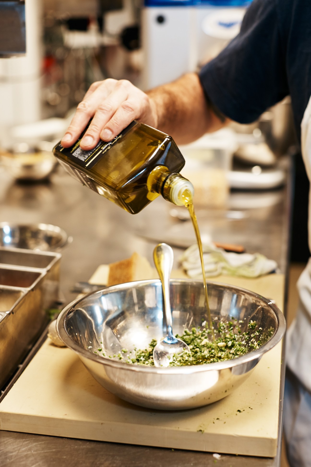 A chef pours olive oil into a metal bowl containing ramp pesto ingredients in a kitchen.
