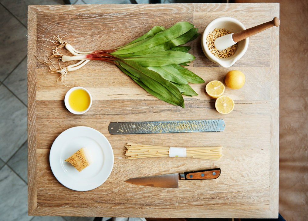 Ramp pesto ingredients arranged on a wooden board including fresh ramps, pine nuts, lemon, olive oil and pasta.