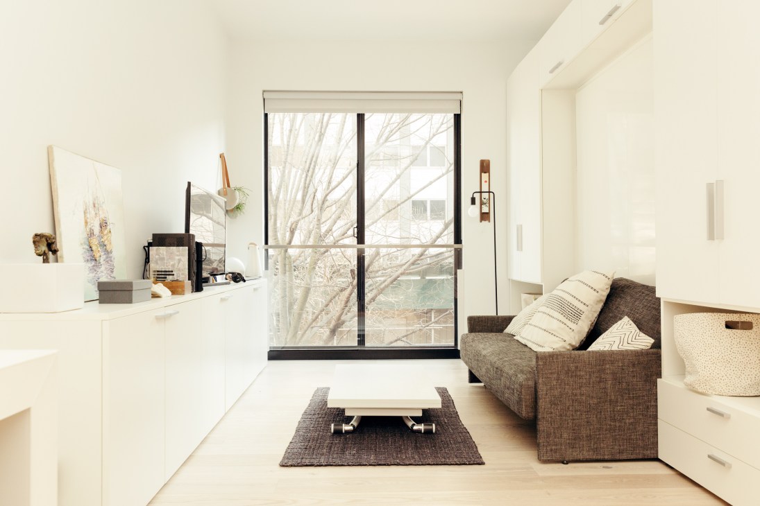 A minimalist micro-apartment at Carmel Place with a brown armchair, height-adjustable coffee table, and large windows overlooking bare trees.