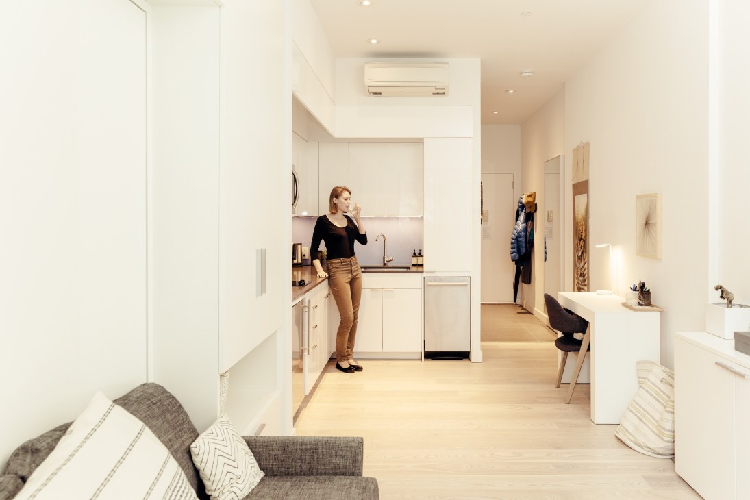 Modern studio kitchen in Carmel Place with white cabinetry, a woman in the foreground, and open-plan living space beyond