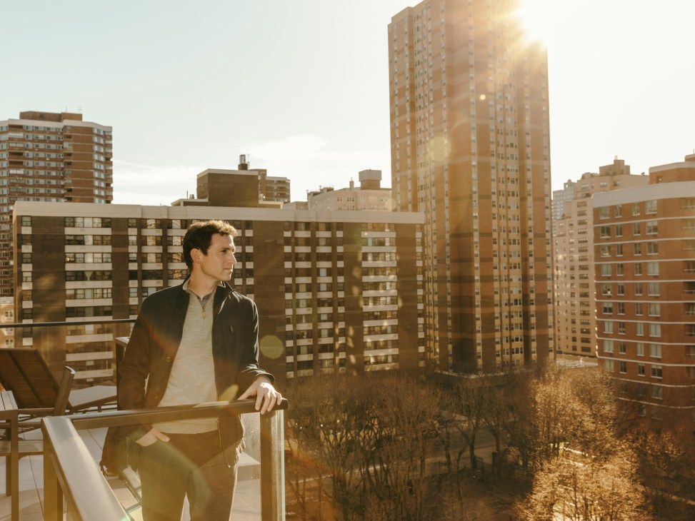 Man standing on a communal roof deck overlooking a cityscape of residential buildings at sunset.