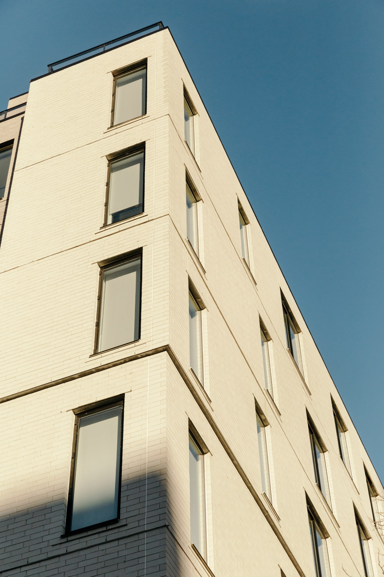 Corner view of Carmel Place, a cream-colored brick building with regularly spaced windows and black trim against a clear blue sky.
