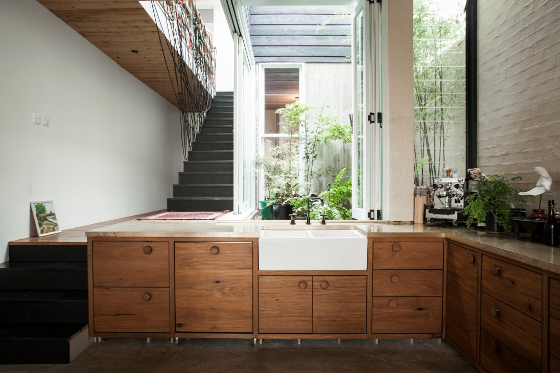 Modern kitchen with wooden cabinetry, white farmhouse sink, and glass doors opening to a courtyard garden.