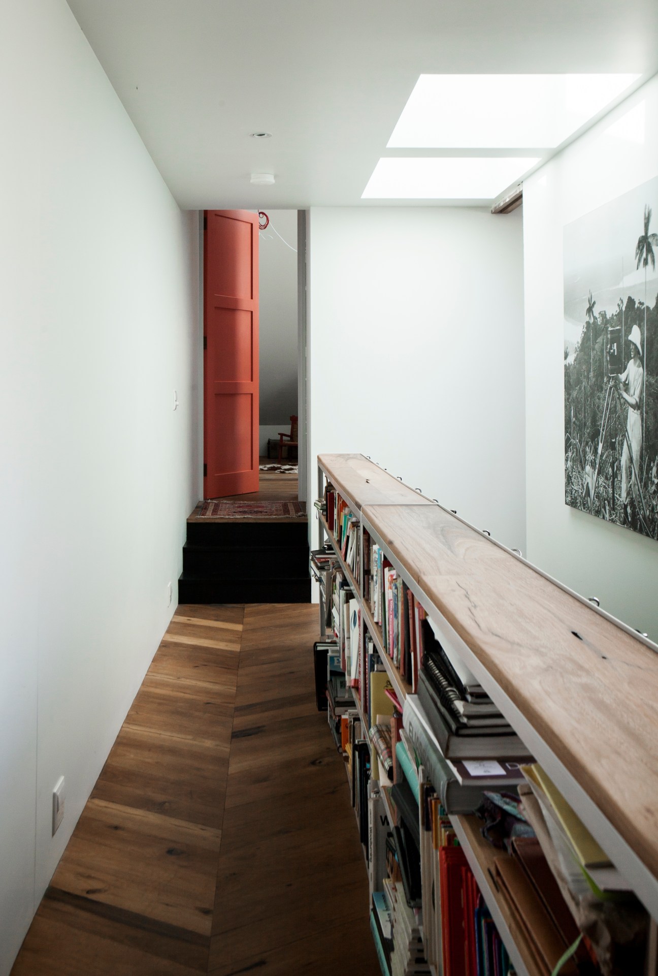 A bright narrow hallway with wooden flooring, white walls, built-in bookshelves, a red door, and skylight windows.