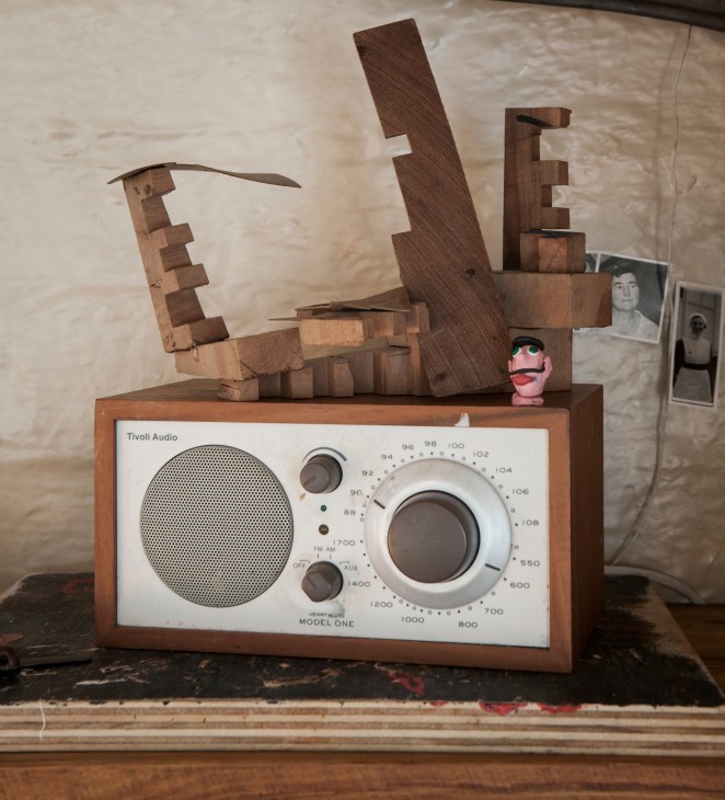 Wooden sculptural art piece displayed atop a vintage Tivoli Audio radio, with photographs visible in the background.