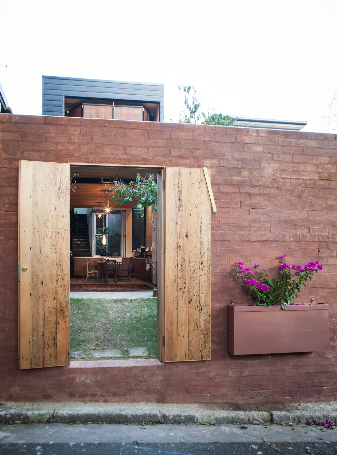 Red brick house exterior with timber-framed glass doors opening to interior, pink flowering plant in matching planter