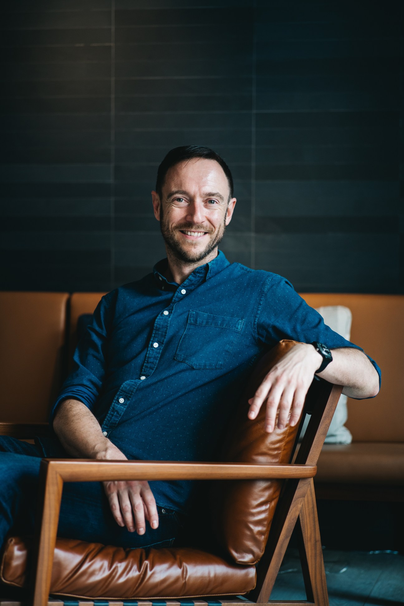 Man in denim shirt sitting on tan leather chair smiling at camera