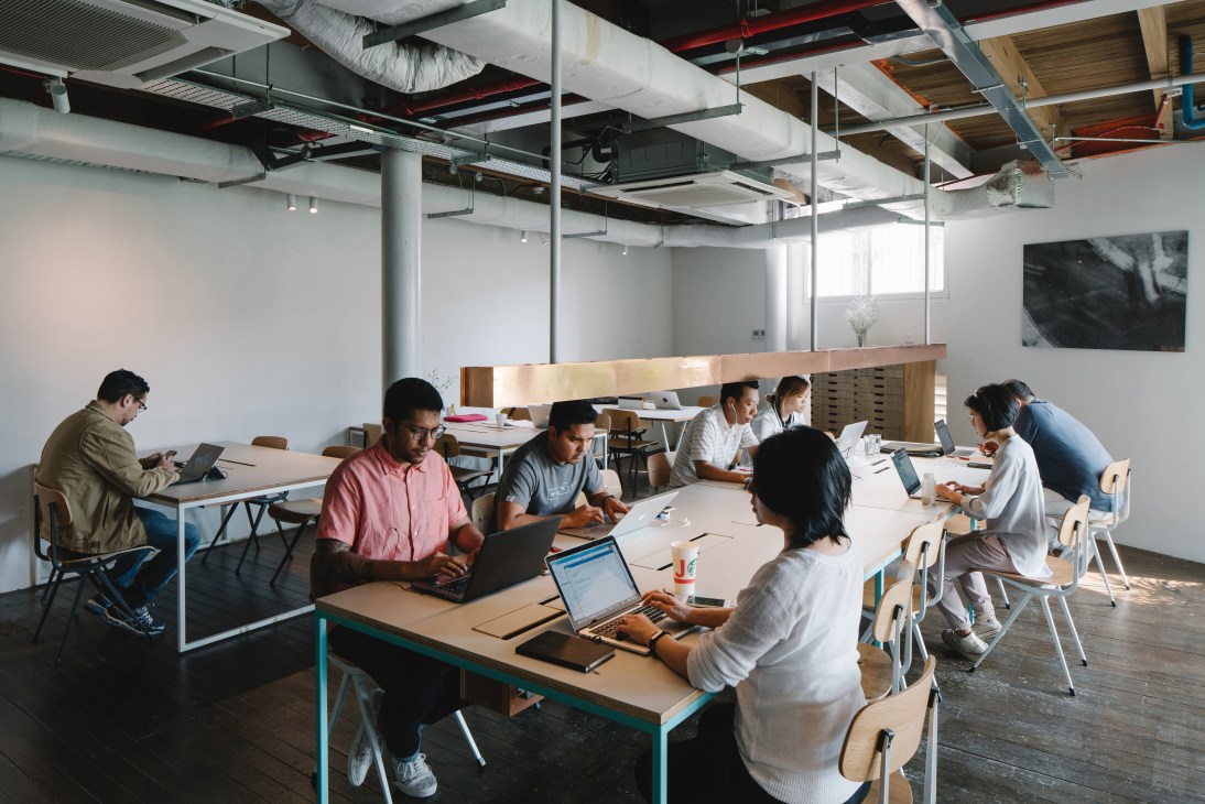 A shared workspace with multiple workers at tables with laptops in a bright, modern industrial loft setting.