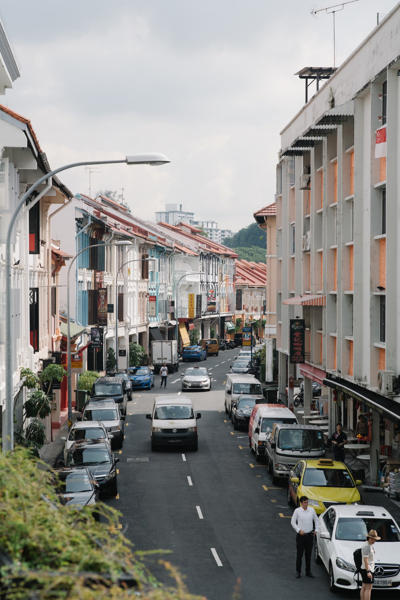 A street view of colorful shophouses in Duxton Hill, Singapore, with parked cars and pedestrians.