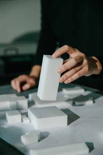 Hand placing white architectural model blocks on a table with scattered miniature building components