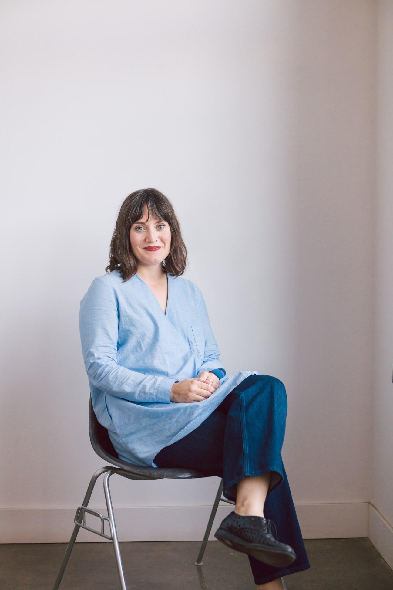 Woman with shoulder-length brown hair wearing light blue tunic sits on black chair against white wall