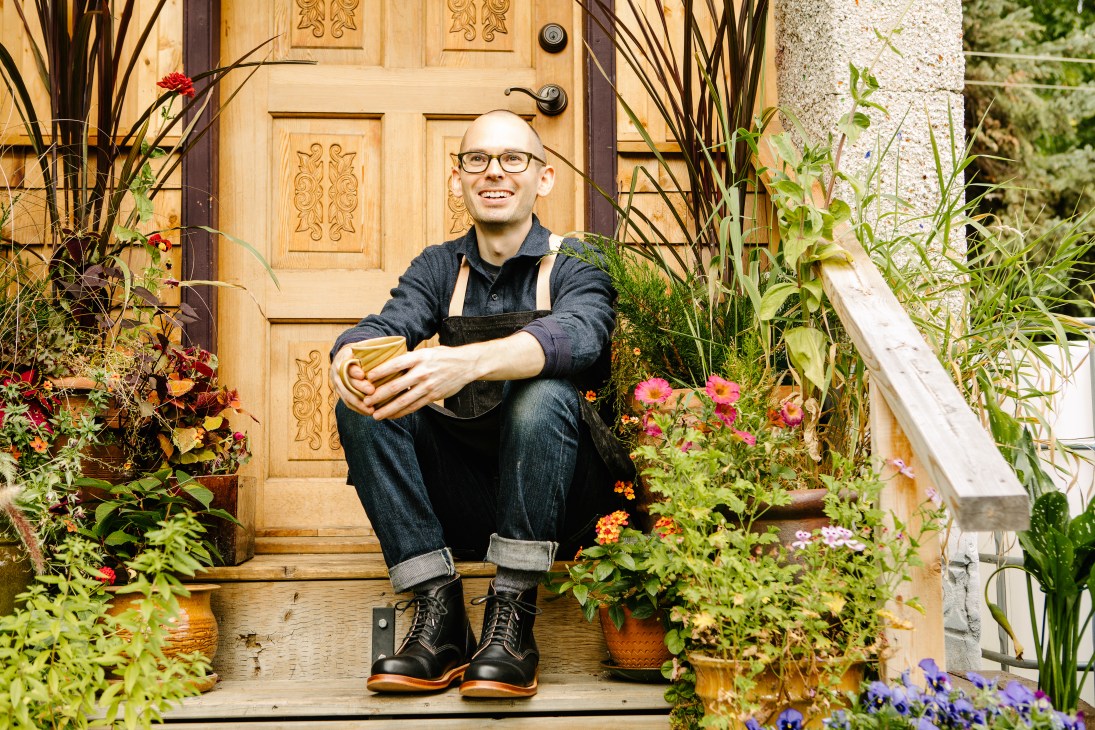 Greg Morgan sits on wooden steps outside ornate door surrounded by potted plants and flowers