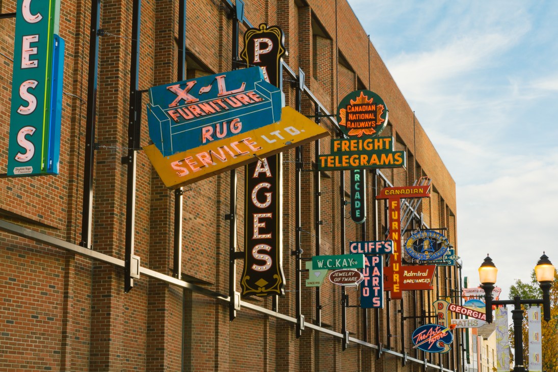 Historic neon signs mounted on a brick wall displaying vintage business names like Pages, X-L Furniture, and Service Co.