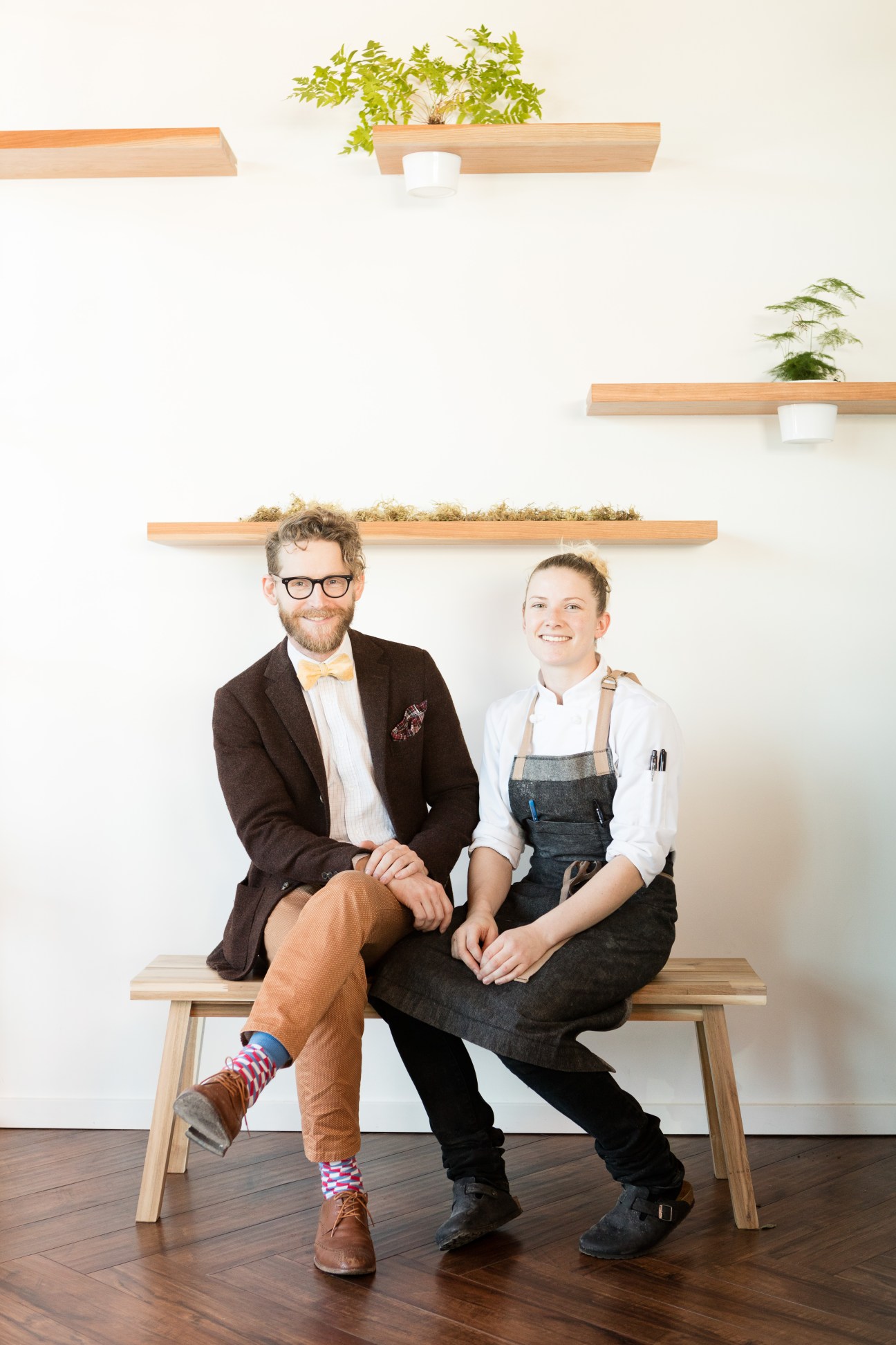 Two people sitting on wooden bench against white wall with floating wooden shelves and plants