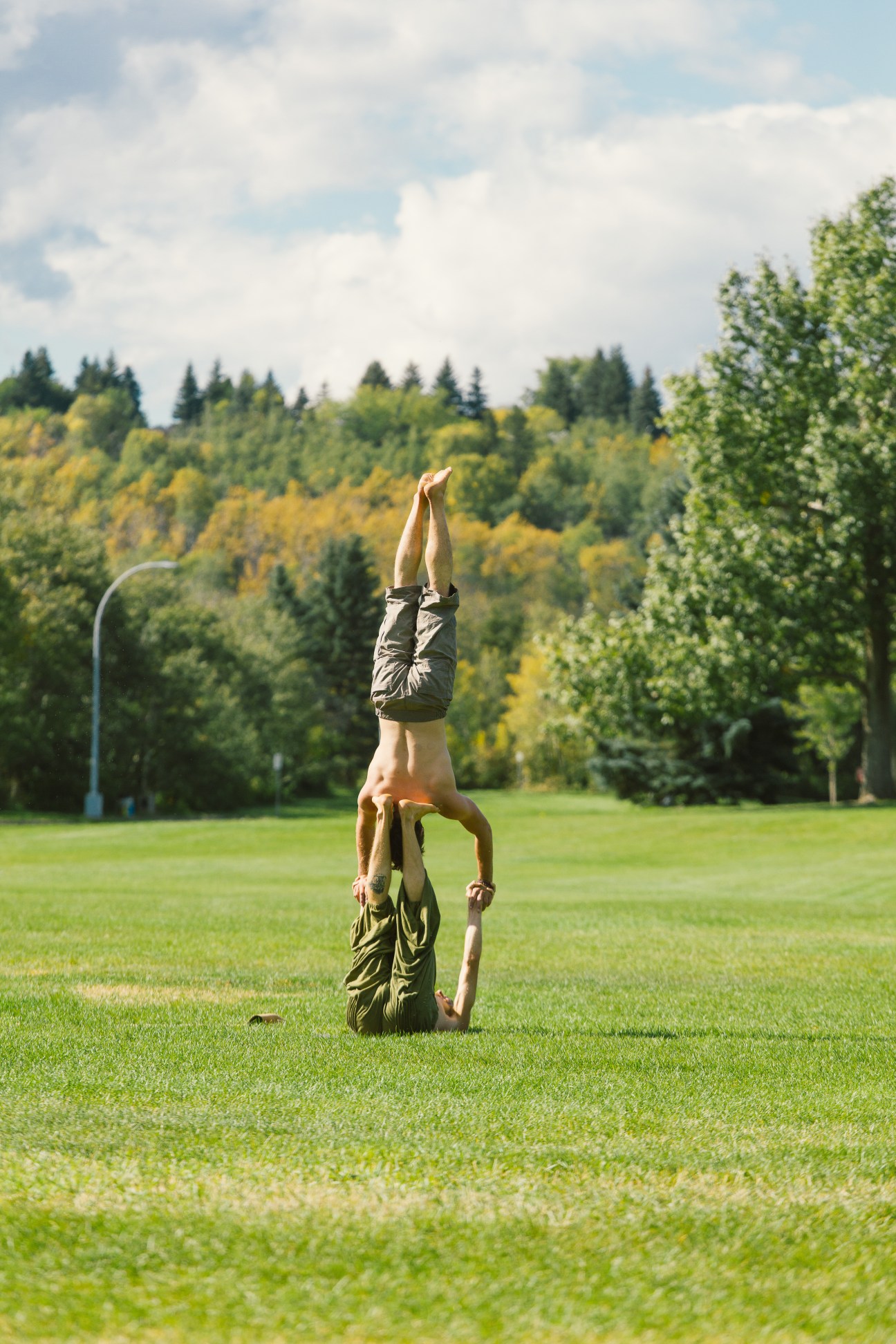 Two people doing acrobatic partner yoga pose on green grass with trees and cloudy sky in background
