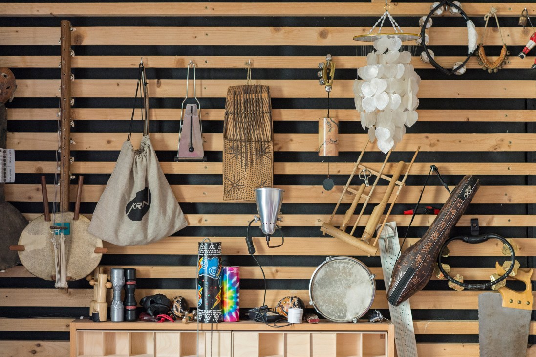 Wooden wall-mounted pegboard displaying various percussion and stringed instruments including marimba, cowbells, tambourines, and hand drums.