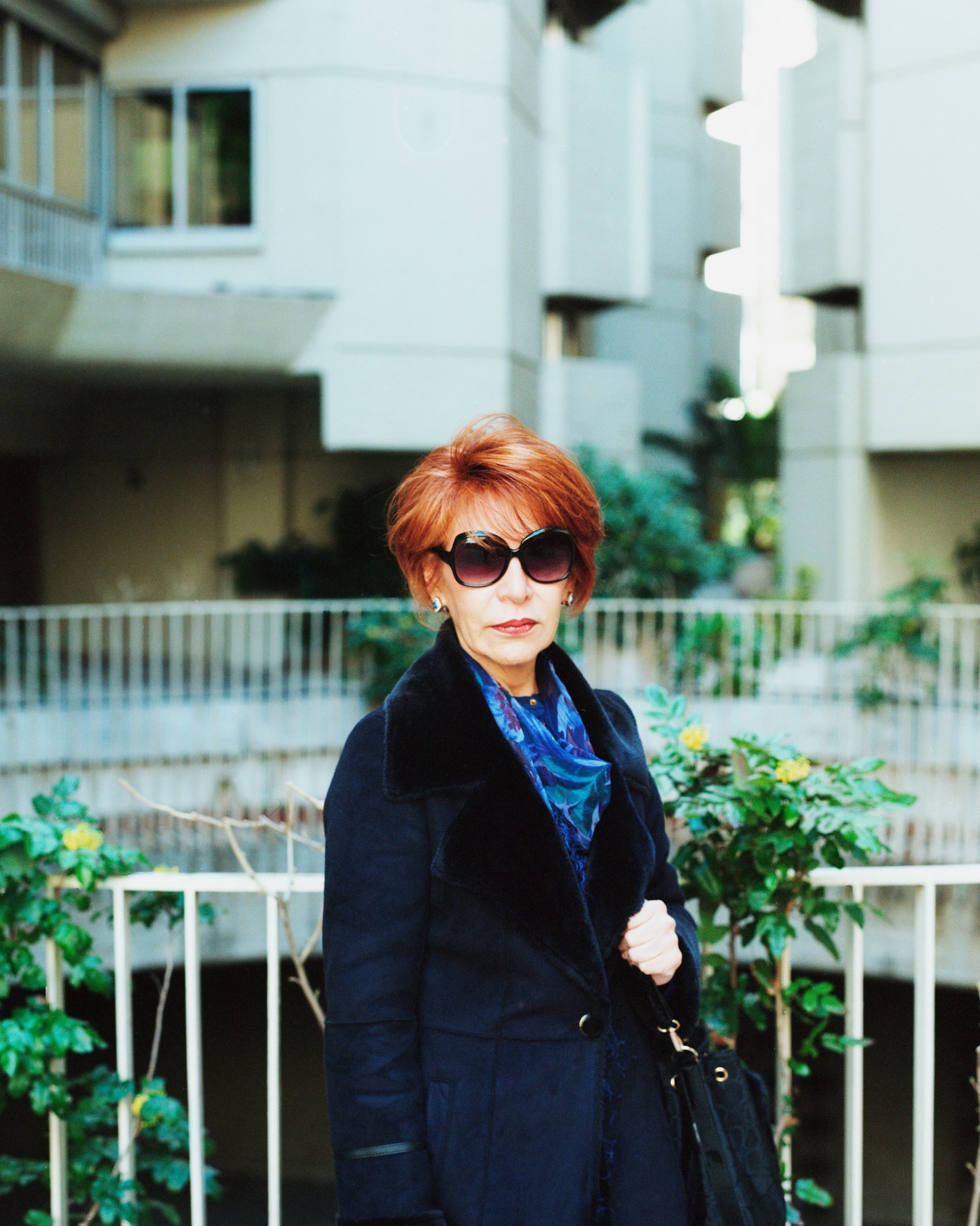 Woman with reddish hair and sunglasses stands in Edificio Princesa's interior courtyard with modernist concrete buildings and climbing plants behind her.