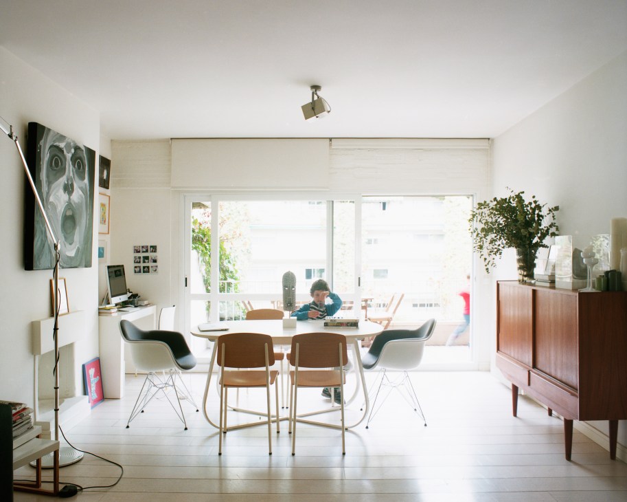 Five-year-old boy sitting at a dining table in a bright, modern apartment with large windows overlooking a building interior.