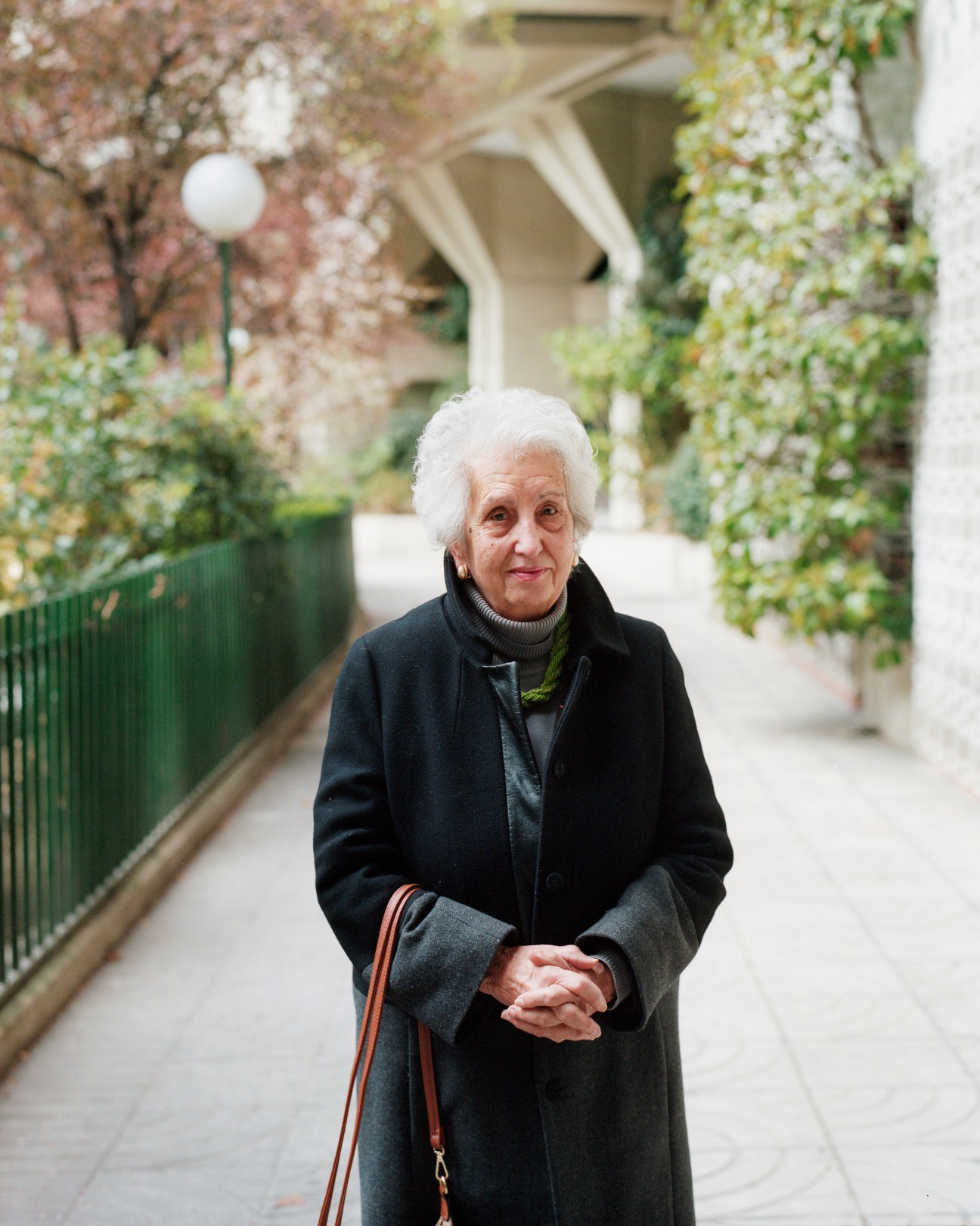 An elderly woman with white hair wearing a black coat walks through a plant-lined courtyard passage.