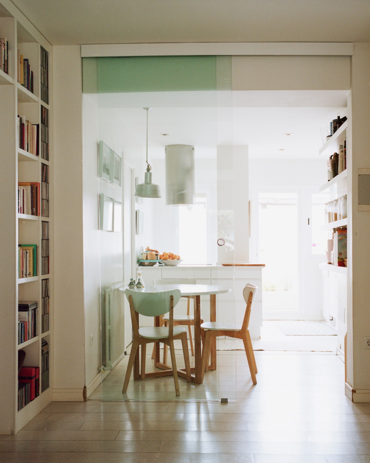 Modern kitchen with glass screen partition, white cabinetry, wooden dining chairs, and built-in bookshelves in a renovated apartment.
