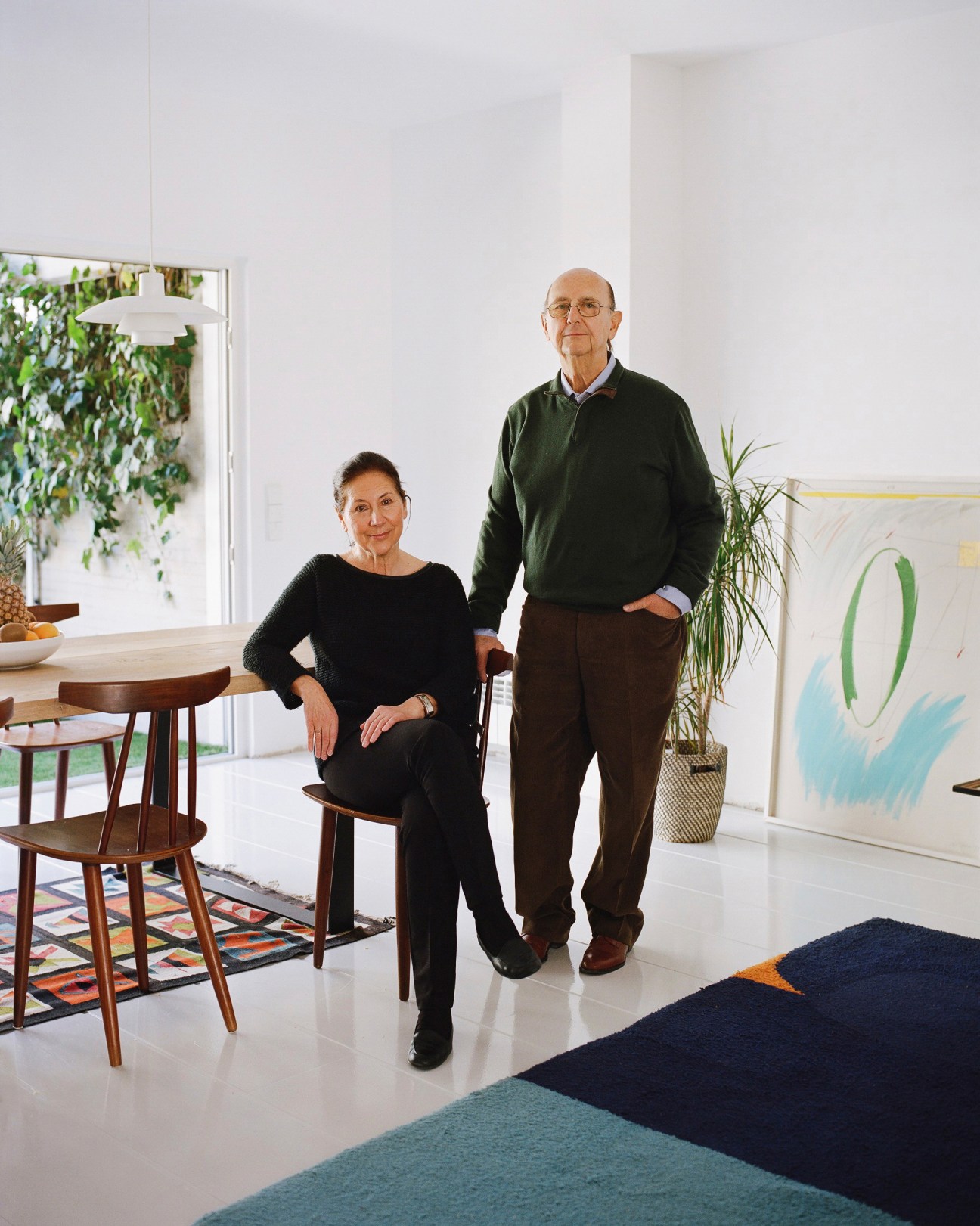 An elderly couple stands in a bright, minimalist apartment with mid-century furniture and hanging plants.