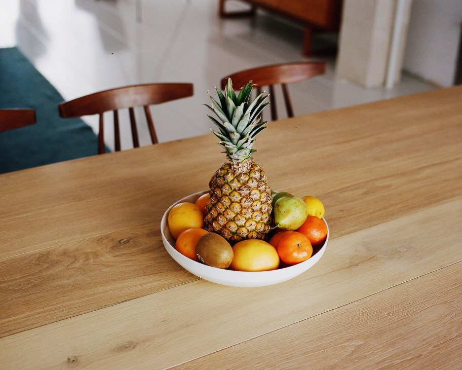 A white bowl of citrus fruits and a pineapple on a wooden dining table with chairs in the background.