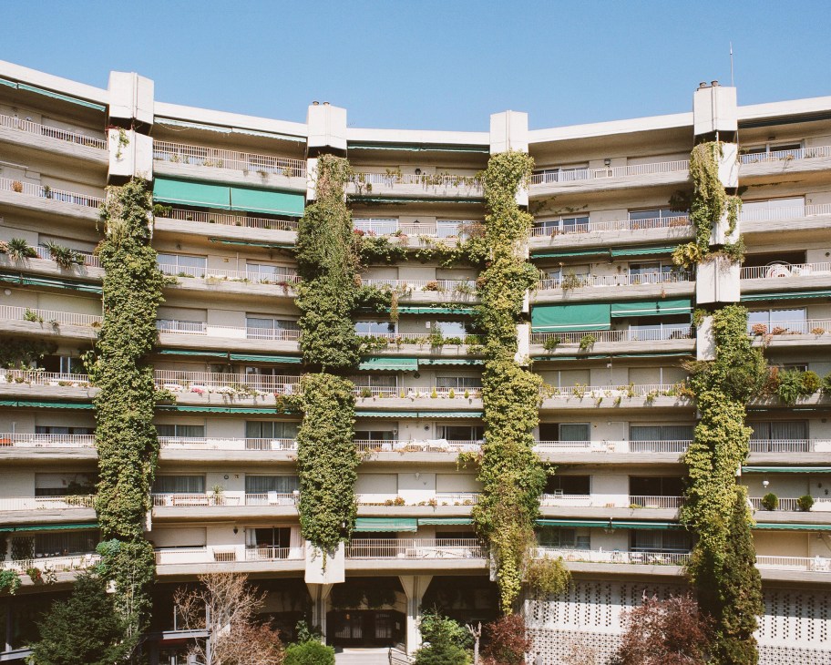 Concrete apartment building façade with ivy-covered vertical columns and multiple balconies facing an interior plaza.