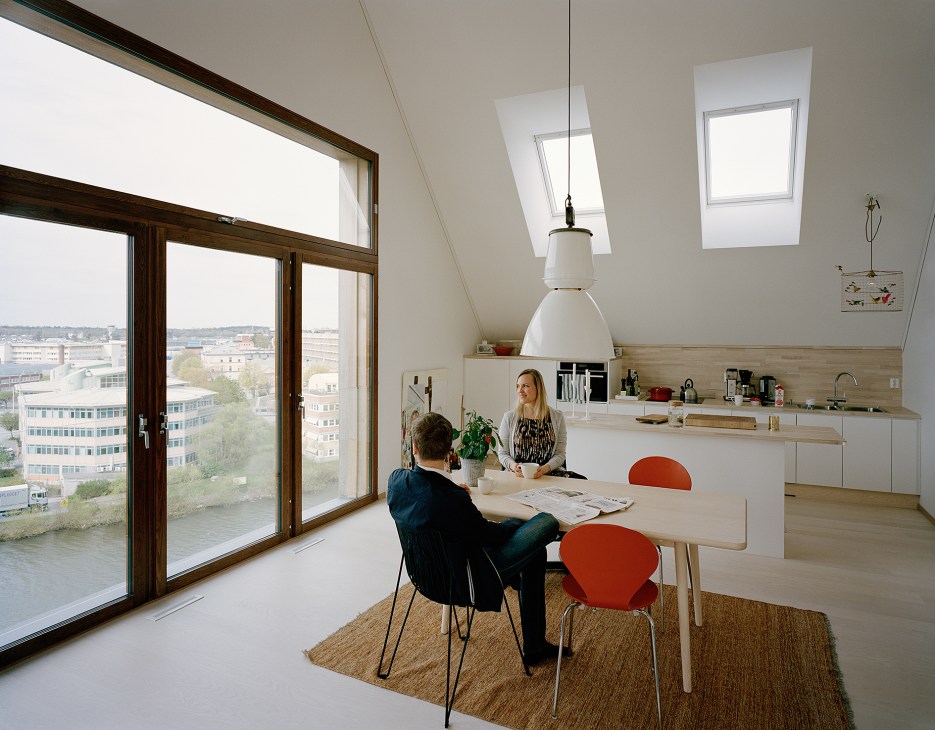 Two people at a dining table in a bright wooden apartment with large windows overlooking Stockholm.