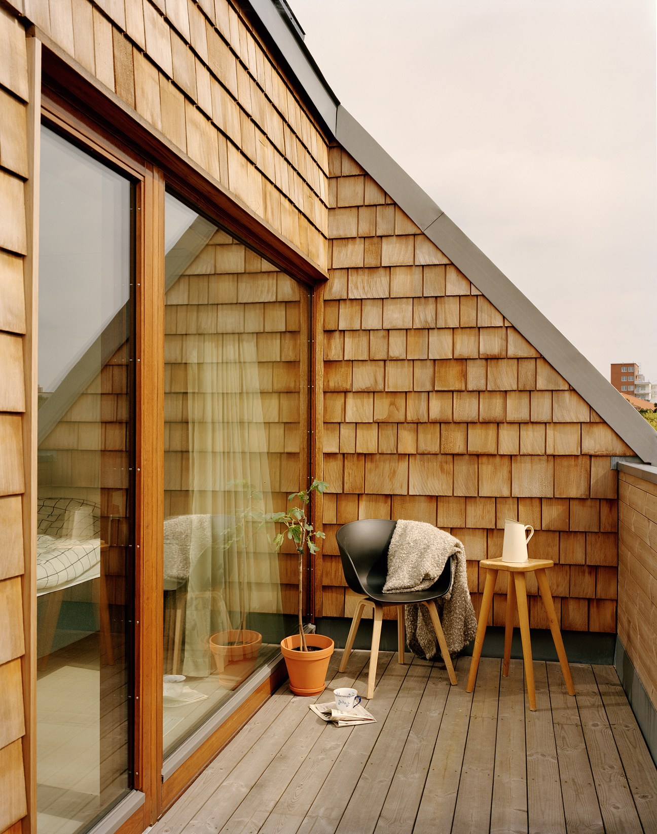 Wooden penthouse balcony with cedar-clad walls, glass doors, wooden deck, black chair, and potted plants.