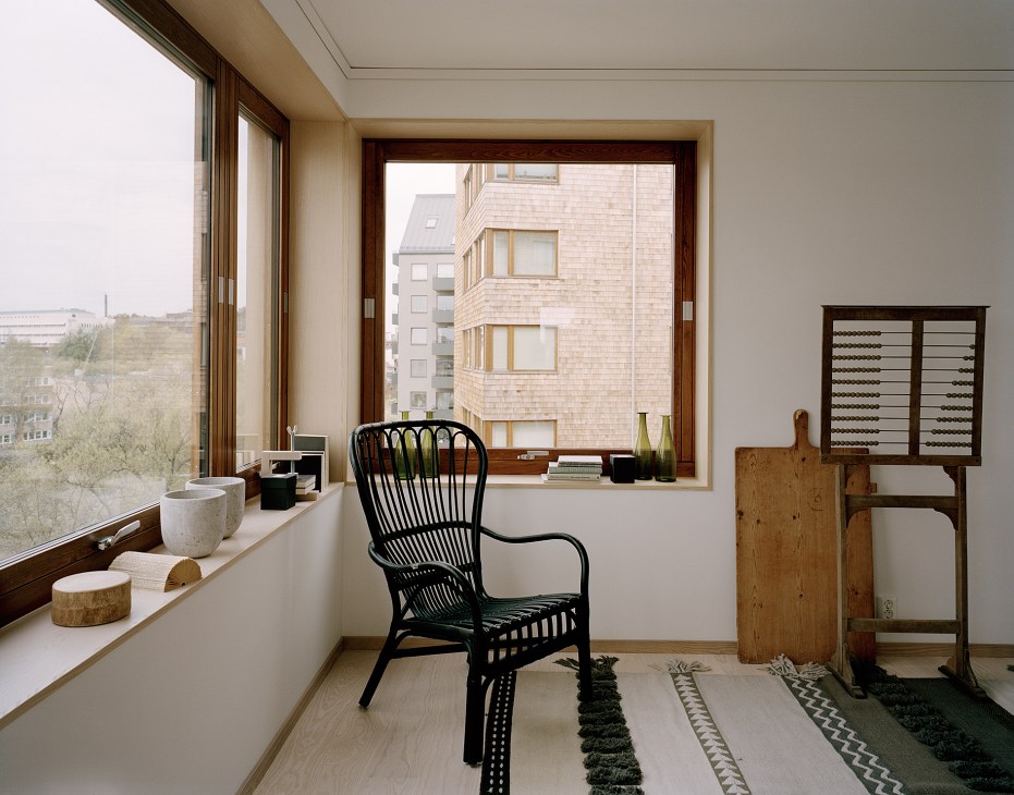 Interior corner of a minimalist apartment with wooden window frames, black wicker chair, and views of wooden apartment buildings outside.