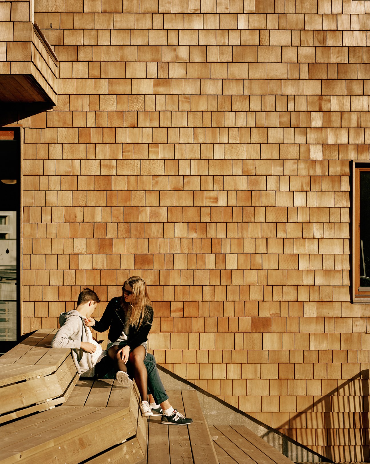 Woman and child sitting on wooden steps against a cedar-clad building facade.