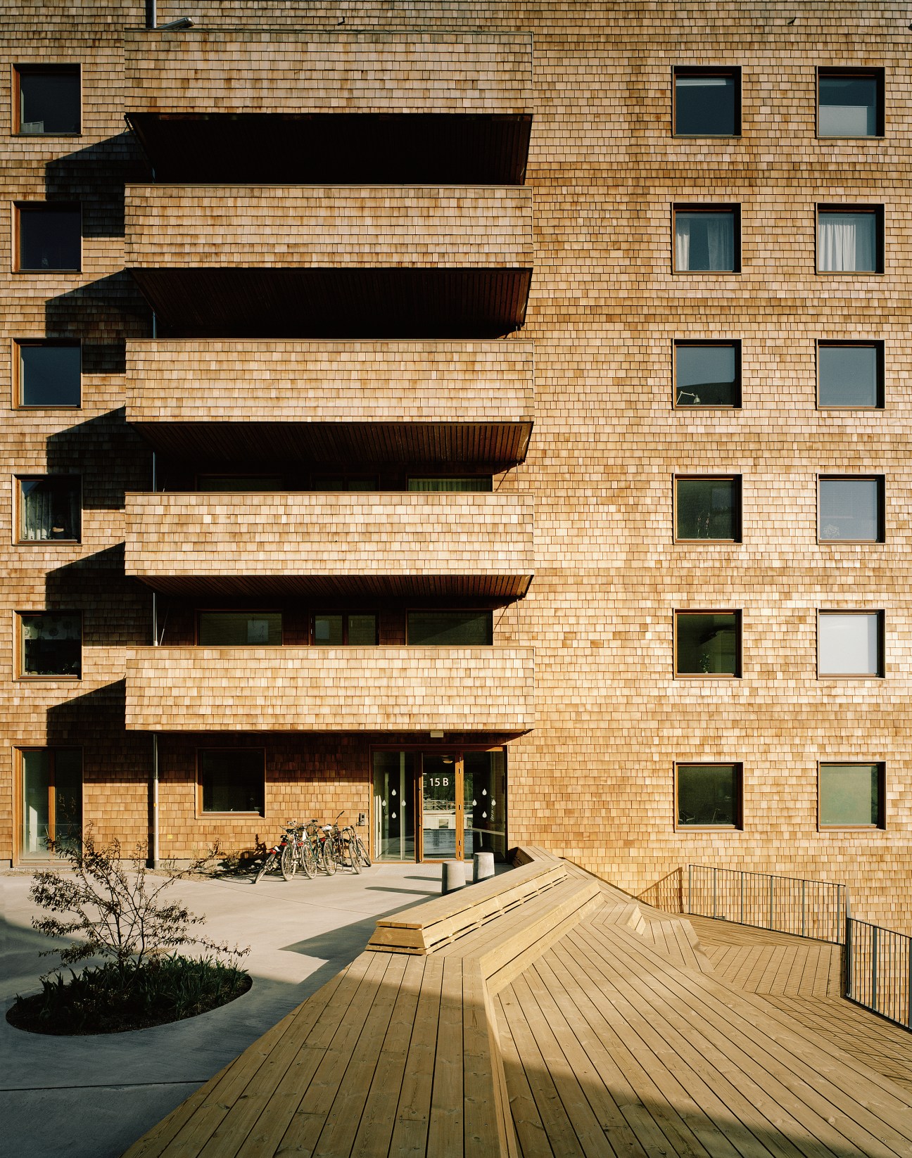 Eight-storey wooden apartment building with cedar-clad facades and dark balconies, featuring a wooden boardwalk entrance.